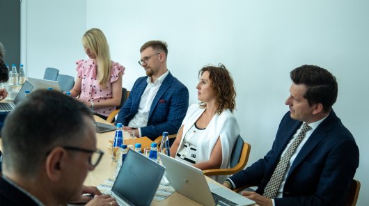 Zdjęcie przedstawia osoby biorące udział w wydarzeniu. Osoby siedzą za stołem i przed nimi stoją laptopy 